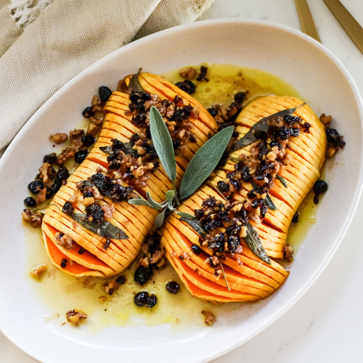 sliced hasselback butternut squash drizzled with melted butter and finished with a crunchy nut-and-dried-fruit topping, garnished with fresh sage on a white platter.