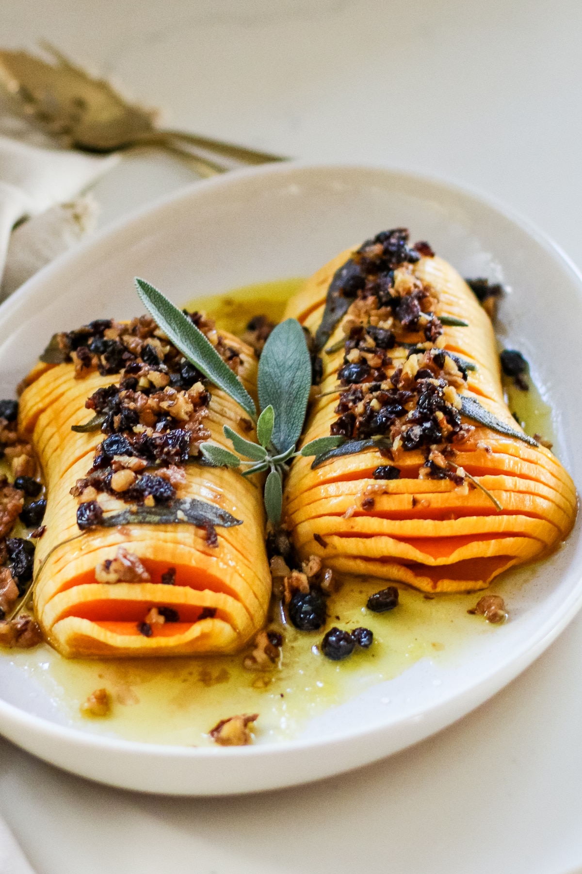 sliced hasselback butternut squash drizzled with melted butter and finished with a crunchy nut-and-dried-fruit topping, garnished with fresh sage on a white platter.