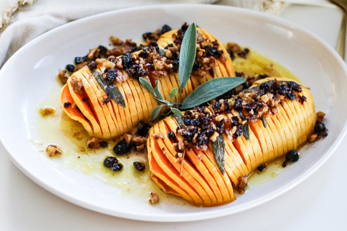 sliced hasselback butternut squash drizzled with melted butter and finished with a crunchy nut-and-dried-fruit topping, garnished with fresh sage on a white platter.