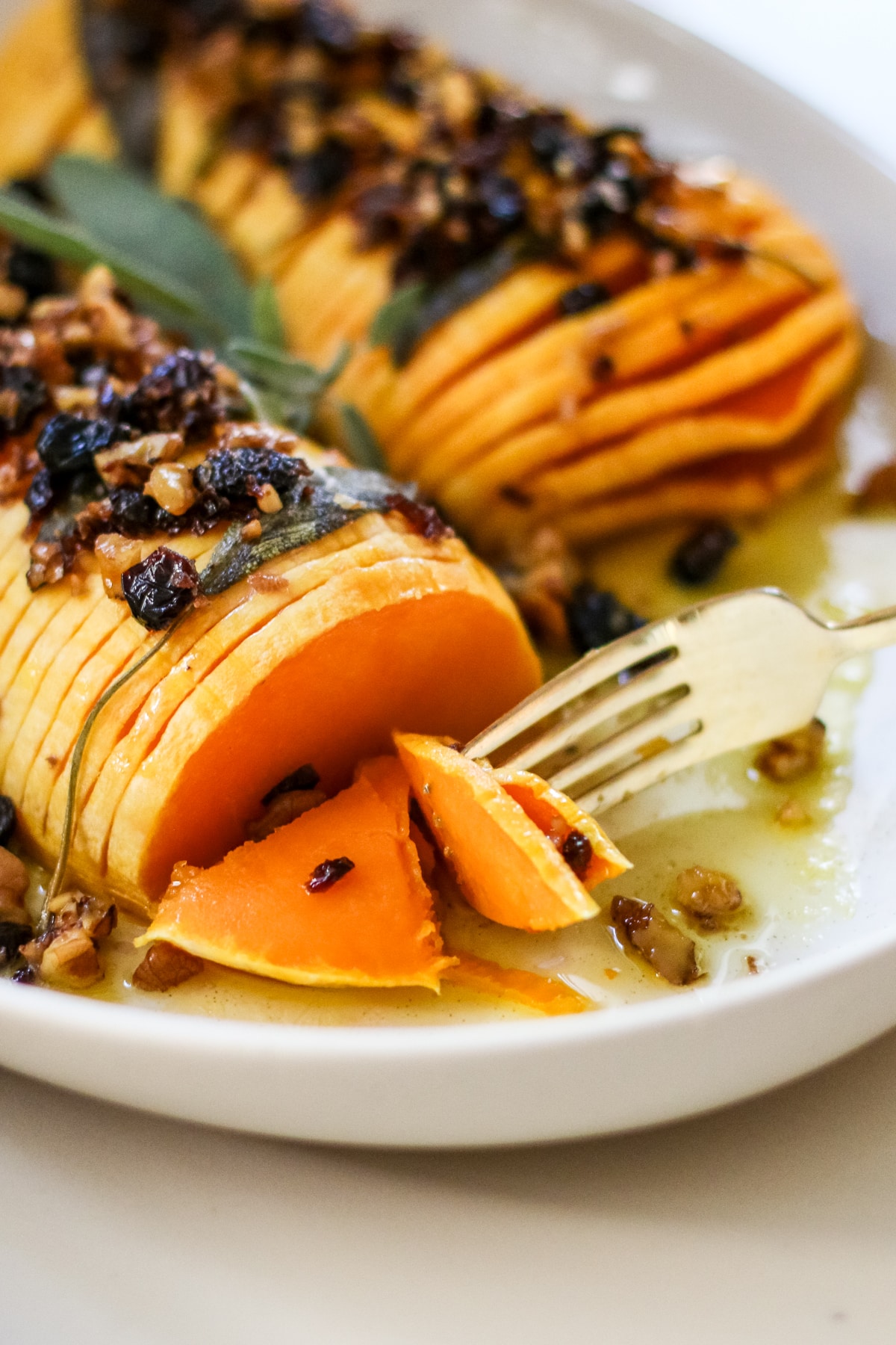 close-up of hasselback butternut squash with a fork lifting a tender slice, topped with browned butter, chopped nuts, dried fruit, and sage on a white plate.