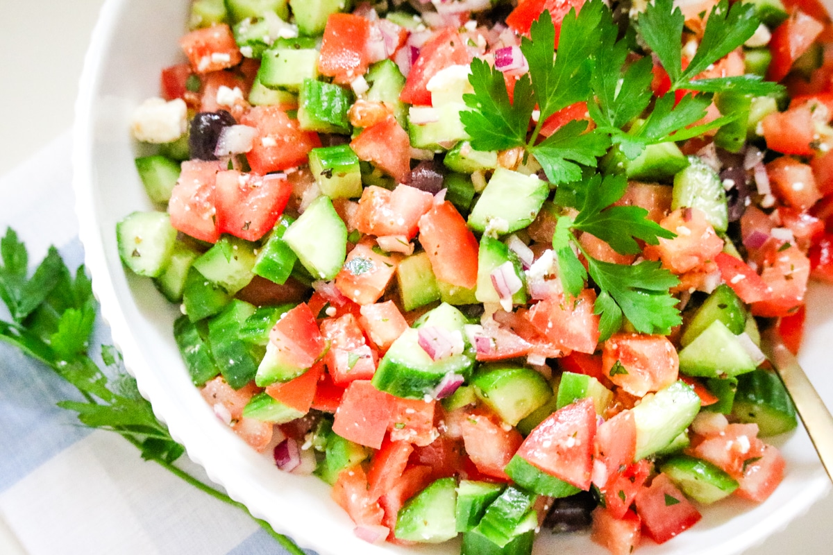 large bowl of Greek salad being tossed with a herb vinaigrette, with the dressing visible coating the cucumbers and tomatoes.