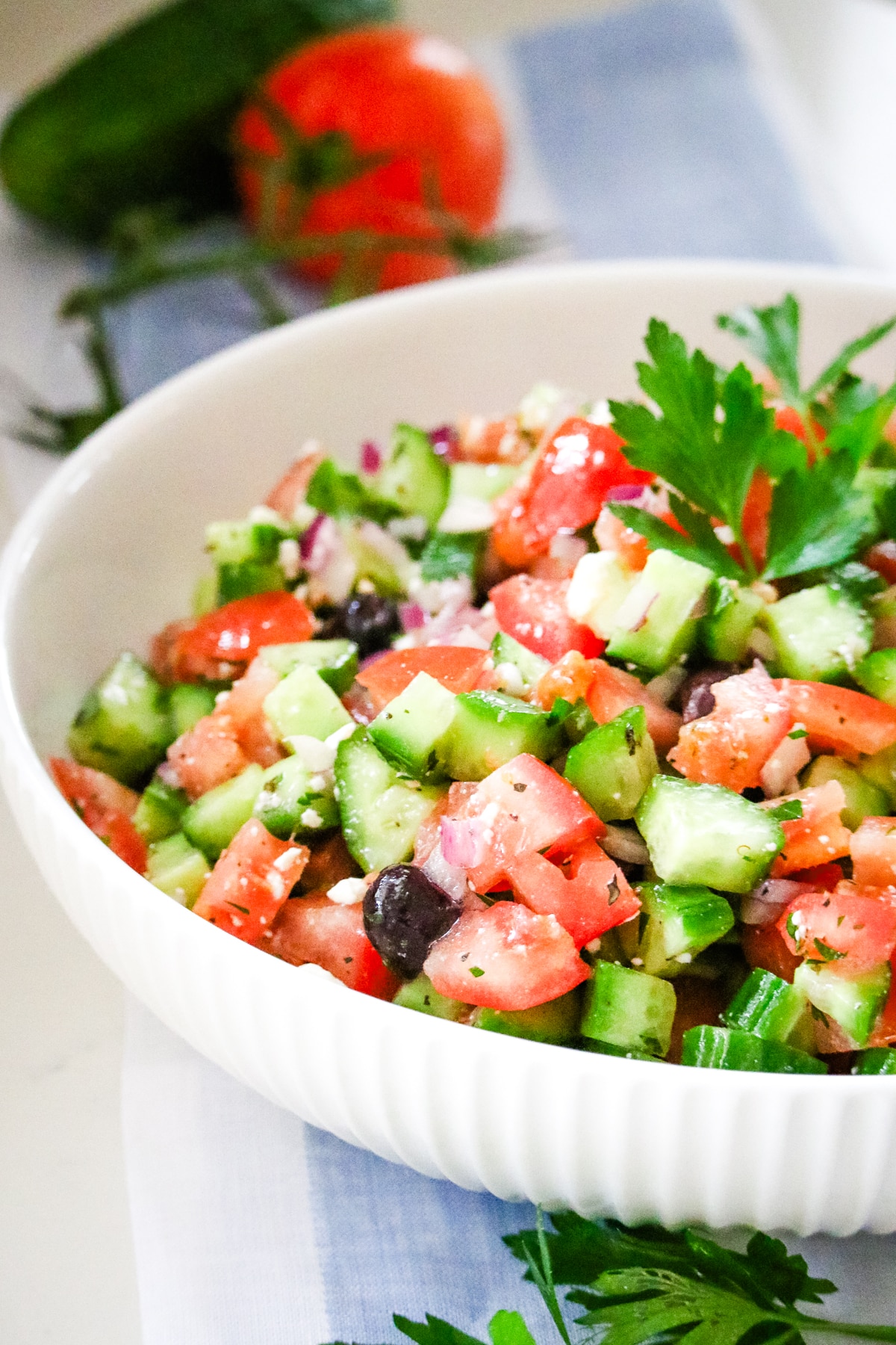 large bowl of Greek salad being tossed with a herb vinaigrette, with the dressing visible coating the cucumbers and tomatoes.