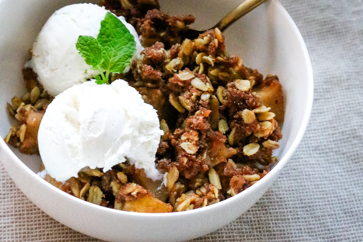 Apple crisp made with fresh-milled flour served in a bowl with a spoon and topped with vanilla ice cream.