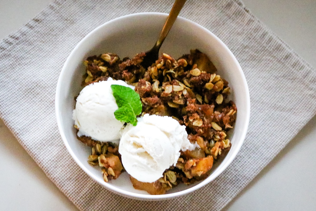 Apple crisp made with fresh-milled flour served in a bowl with a spoon and topped with vanilla ice cream.