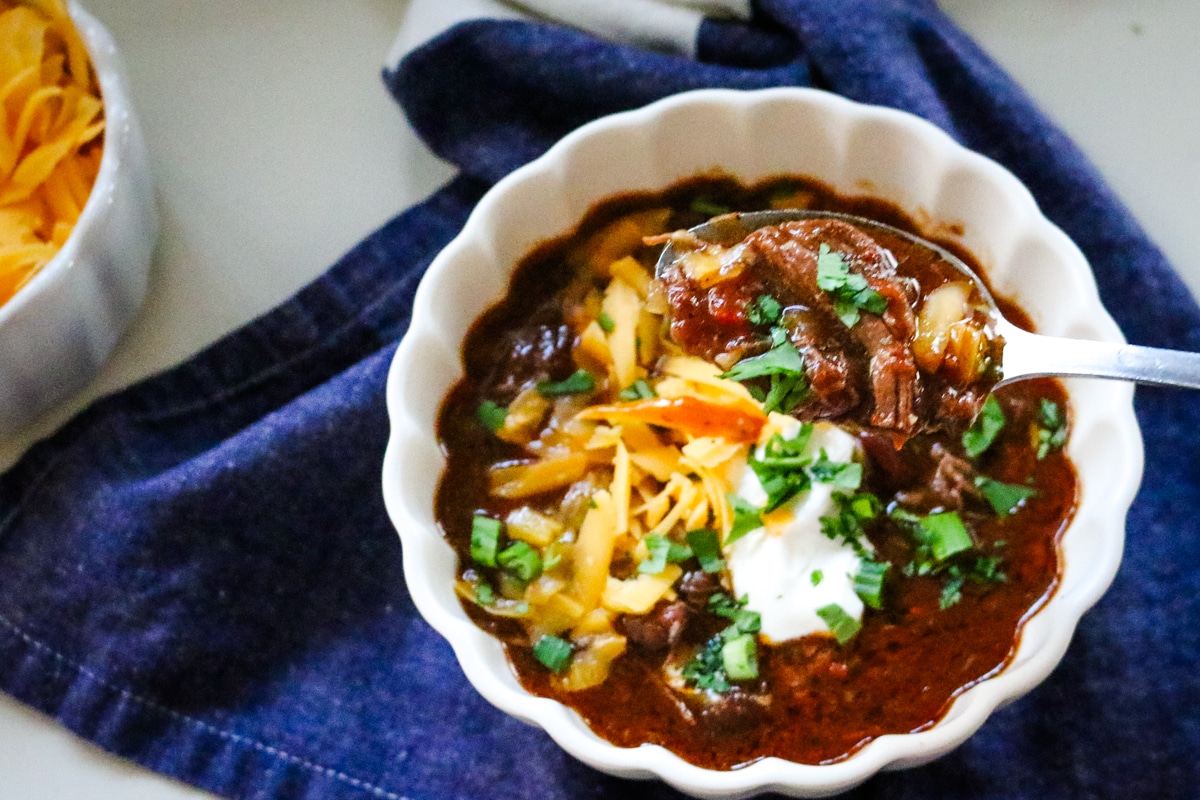 shredded beef chili on a spoon in a white bowl and blue kitchen towel.