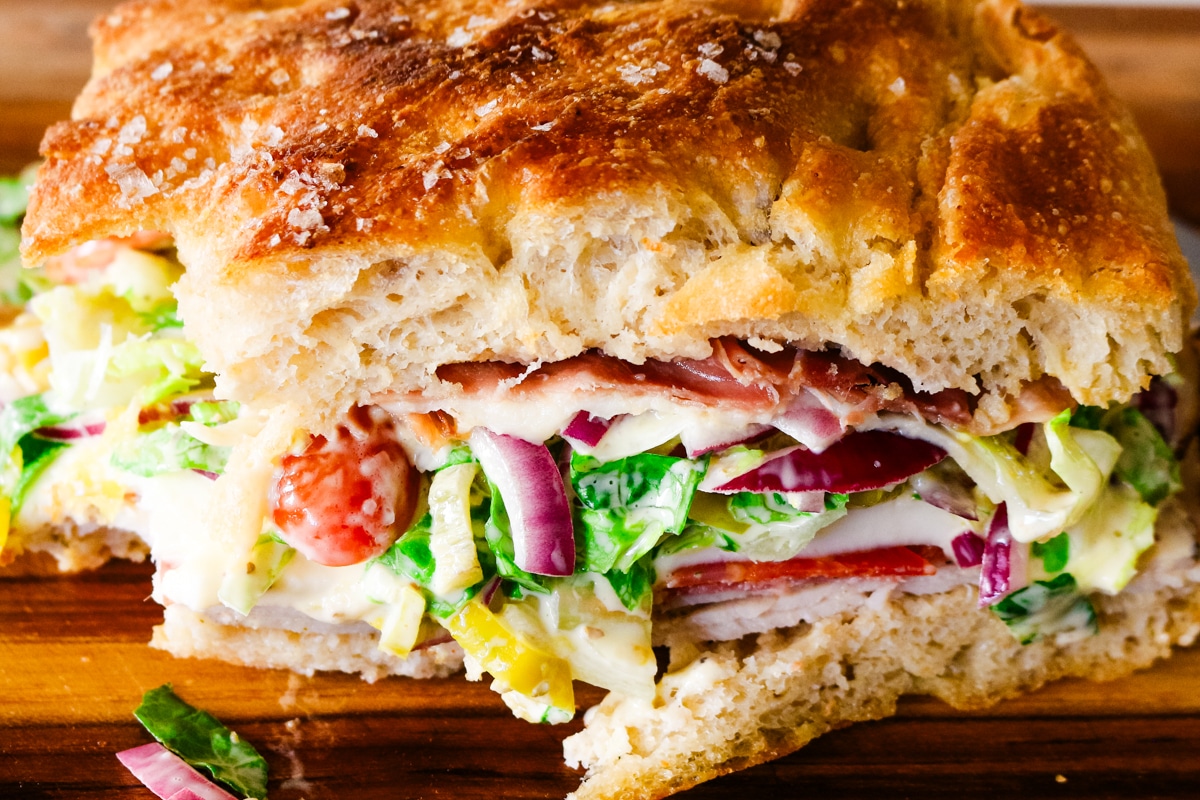 grinder sandwich on a focaccia bread with red onions, deli meats, and shredded cabbage salad.