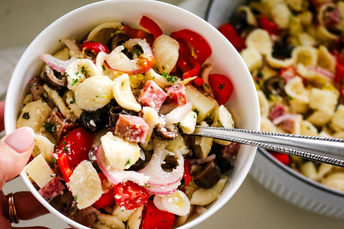 italian pasta salad in a white bowl being held above a bigger bowl of italian pasta salad.
