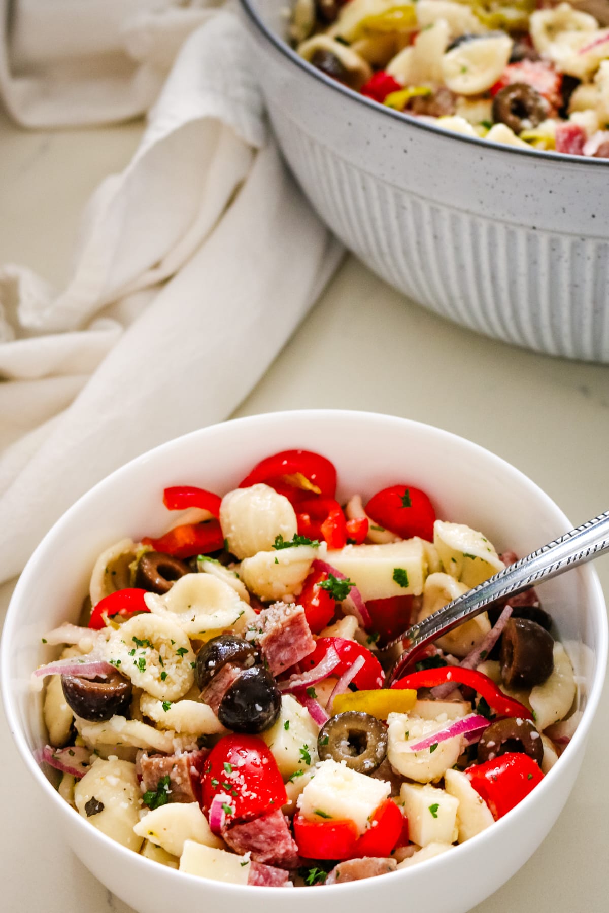 italian pasta salad in a white bowl with a fork and a bigger platter filled with pasta salad on the side.