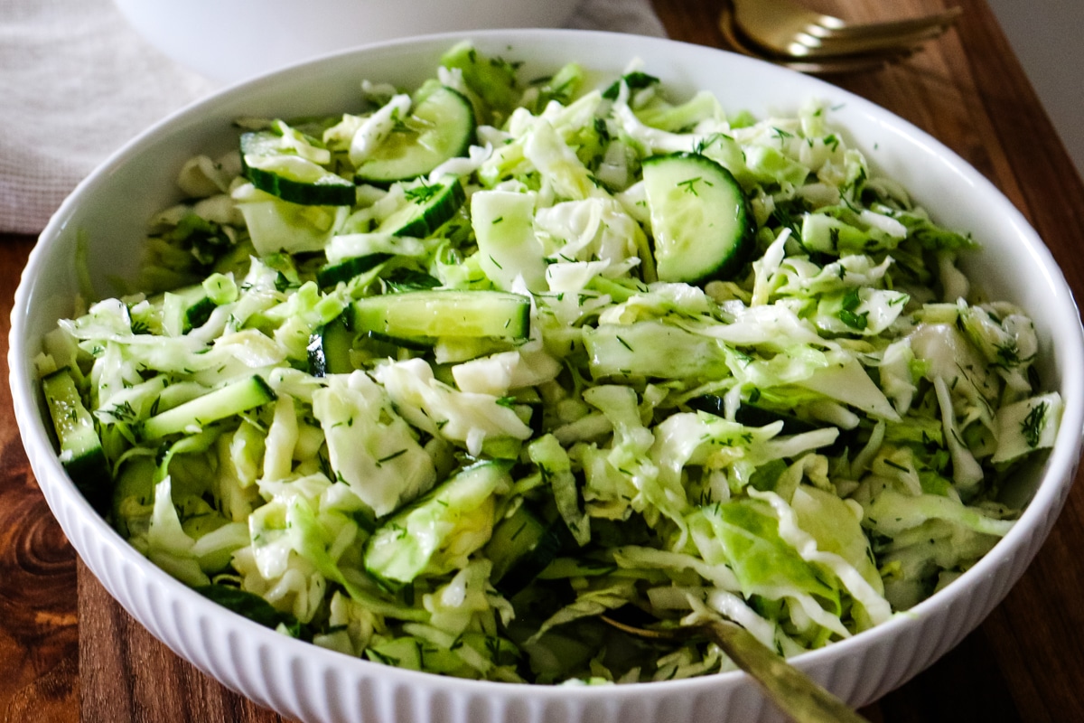 A wide overhead view of a large white bowl filled to the brim with freshly tossed cucumber cabbage salad and dill, resting on a wooden surface next to a fork.