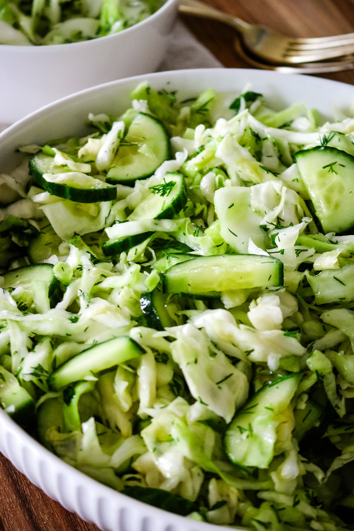 A tight, angled close up of a white serving bowl filled with cucumber cabbage salad and fresh dill, with a smaller bowl and a fork resting on a wooden surface partially visible in the background.