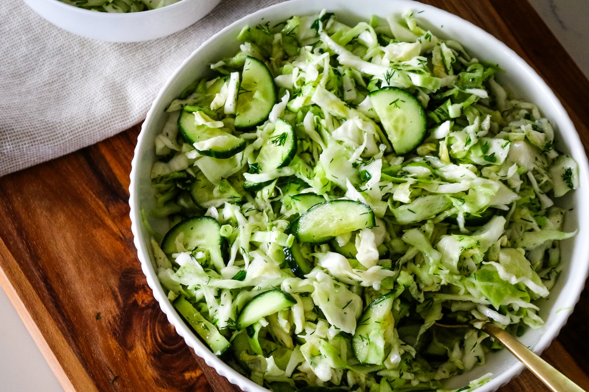 A large white serving bowl filled with freshly tossed cucumber cabbage salad, garnished with fresh dill, sitting on a wooden cutting board with a serving spoon resting inside.
