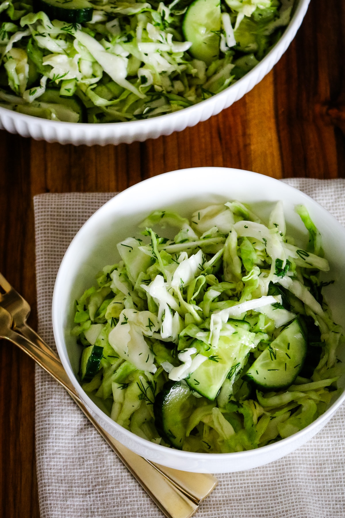 A small bowl filled with freshly tossed cucumber cabbage salad, garnished with fresh dill, sitting on a cloth napkin alongside a fork, with a larger serving bowl of the same salad in the background on a wooden cutting board.