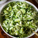 A close up, square shot of a large textured ceramic bowl filled with freshly tossed cucumber cabbage salad and fresh dill, resting on a wooden cutting board with a metal serving spoon tucked inside.