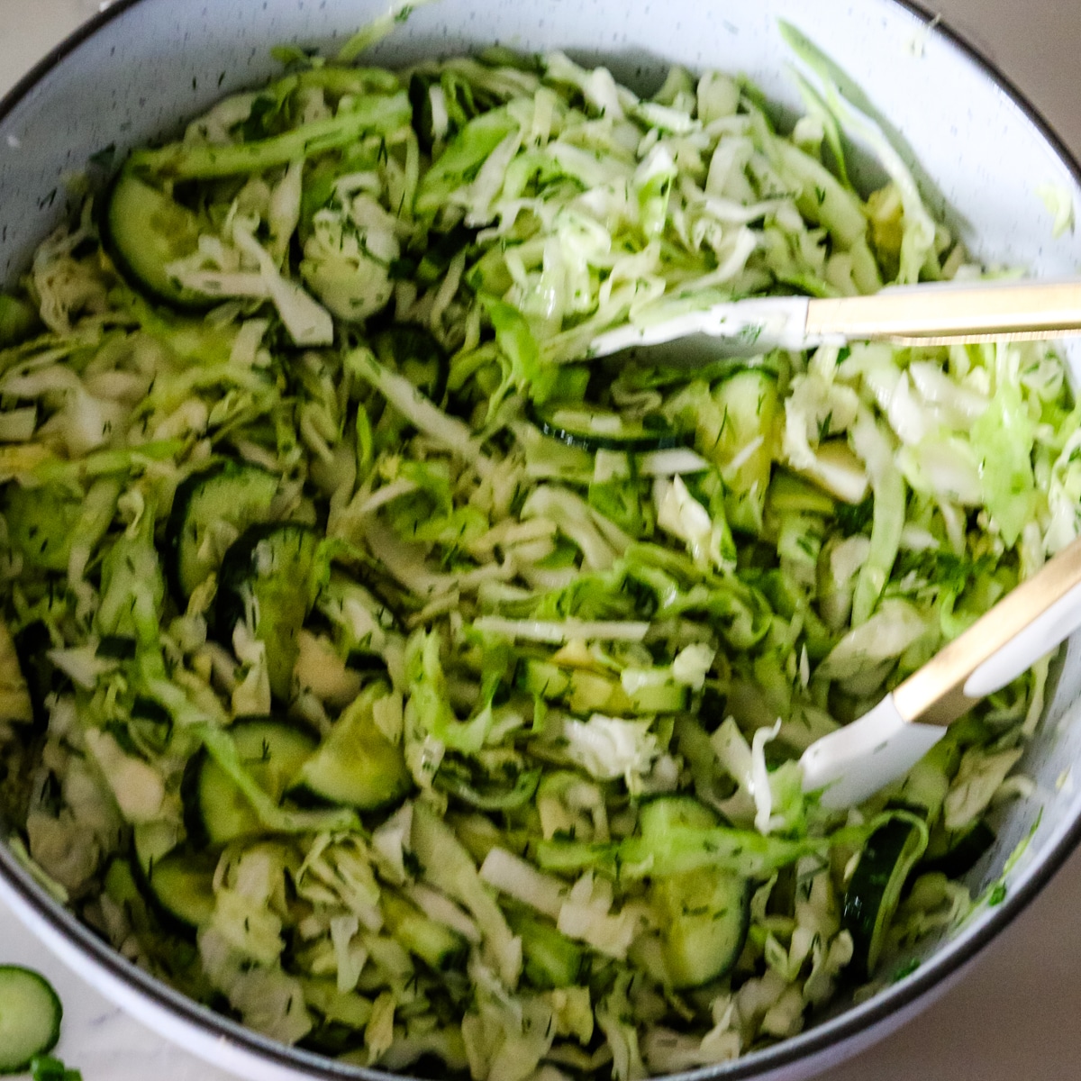 Tossing shredded cabbage, sliced cucumbers, and chopped dill with salad servers in a large mixing bowl to combine the ingredients.