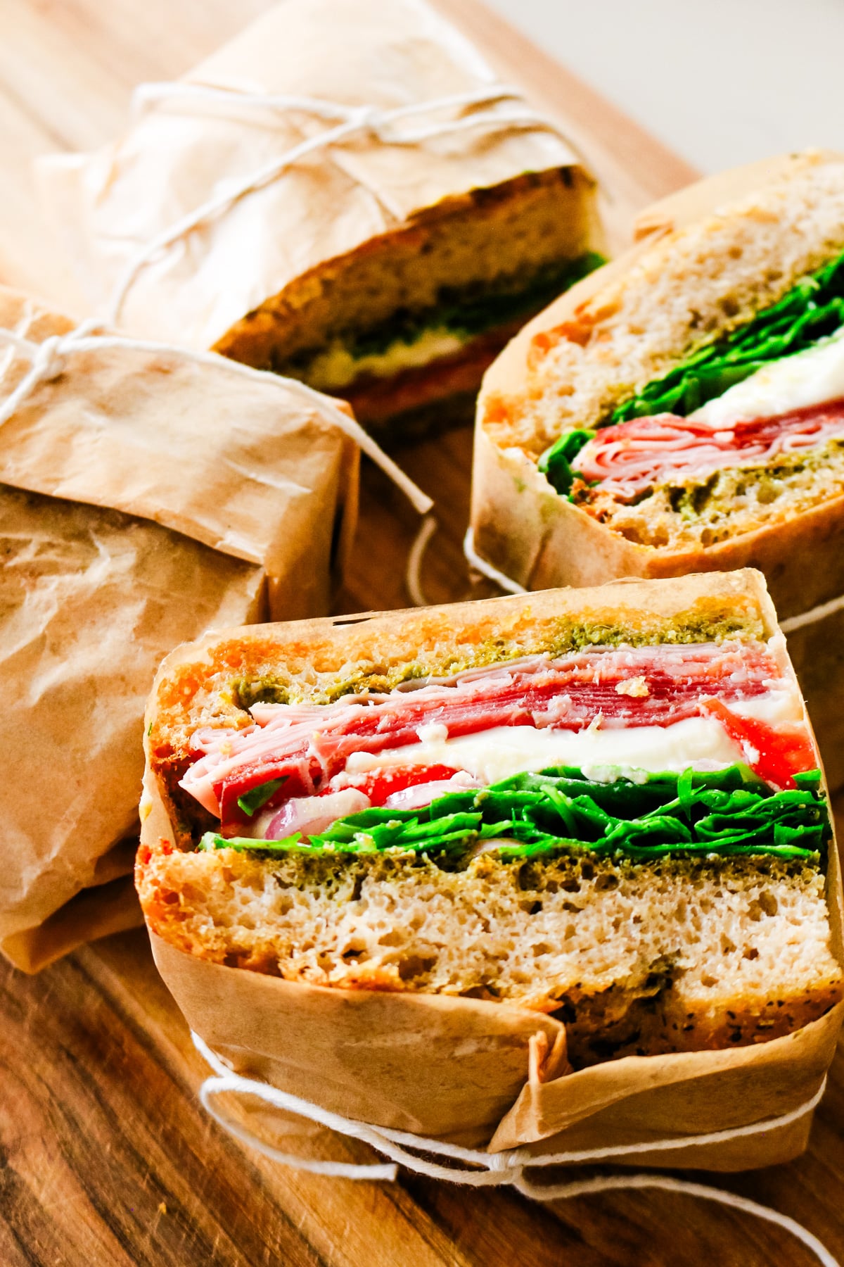 Halved Italian sandwiches wrapped in brown parchment paper showing rich layers of cured meats, fresh mozzarella, pesto, and greens served on a wooden board.