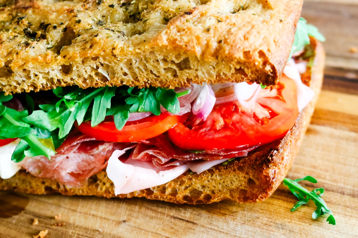 Close-up of golden, seasoned focaccia bread packed with fresh arugula, ripe tomato slices, sliced shallots, and delicate layers of deli meats.