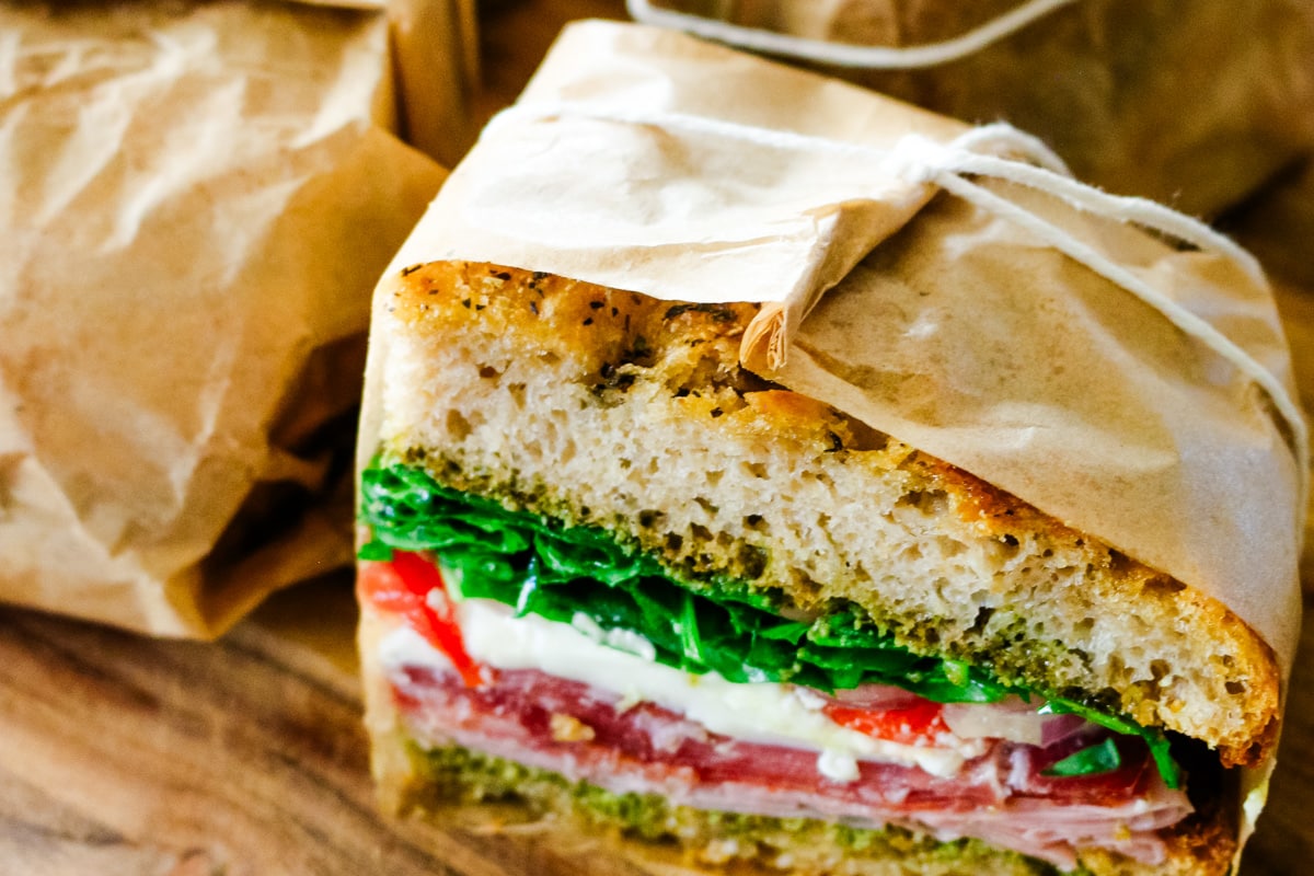 Close up of parchment-wrapped Italian sandwich halves tied with twine, showing layers of pesto, cured meats, and fresh mozzarella on artisan bread.