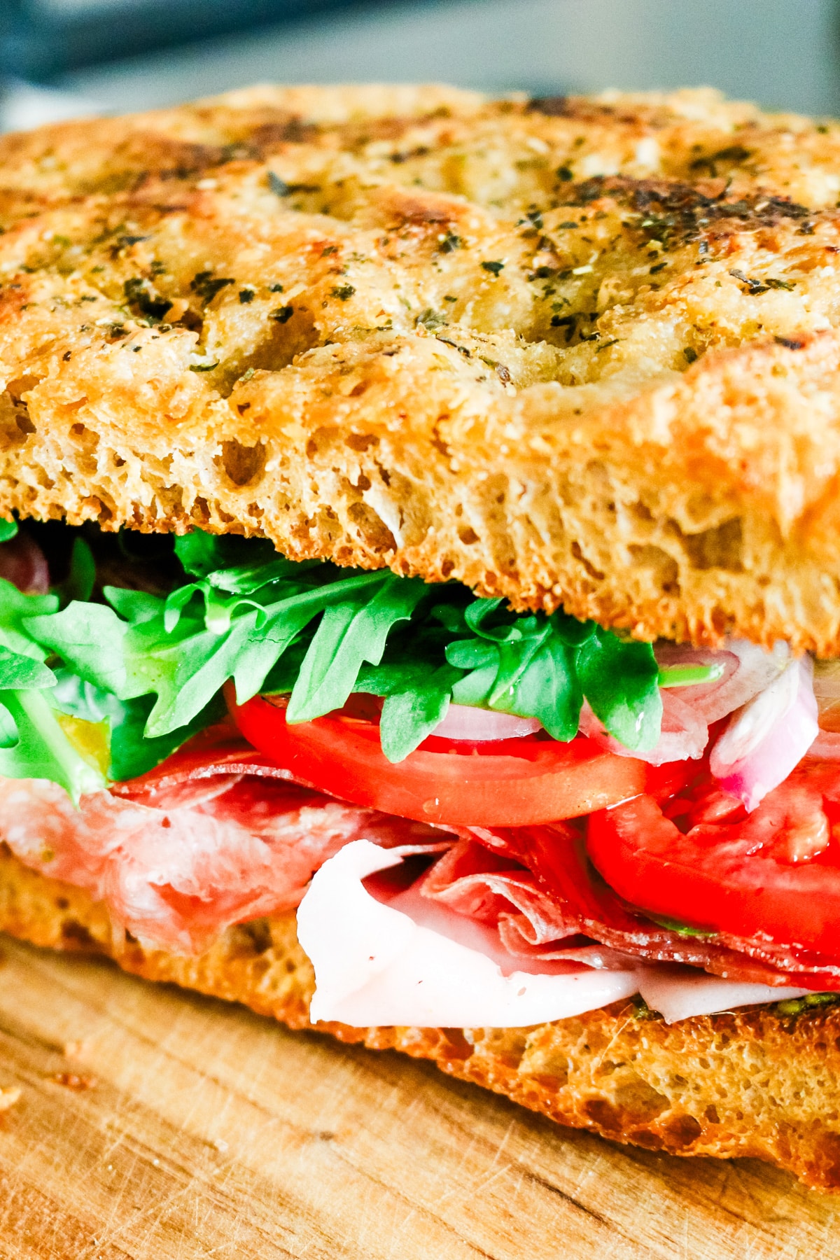 Close-up view of an Italian sandwich made with herb focaccia bread, cured meats, fresh tomatoes, and arugula.