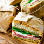 Parchment-wrapped Italian sandwich halves tied with twine, showing layers of pesto, cured meats, and fresh mozzarella on artisan bread.