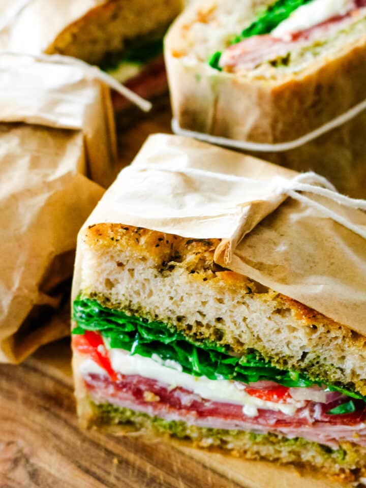 Parchment-wrapped Italian sandwich halves tied with twine, showing layers of pesto, cured meats, and fresh mozzarella on artisan bread.