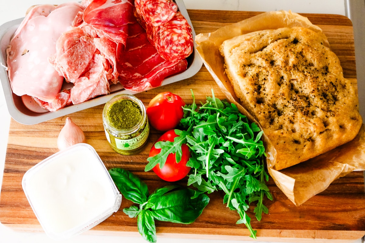 An overhead view of Italian sandwich ingredients arranged on a wooden board, including a tray of sliced cured meats, a loaf of focaccia bread, fresh tomatoes, arugula, basil, a jar of pesto, fresh mozzarella, and a shallot.