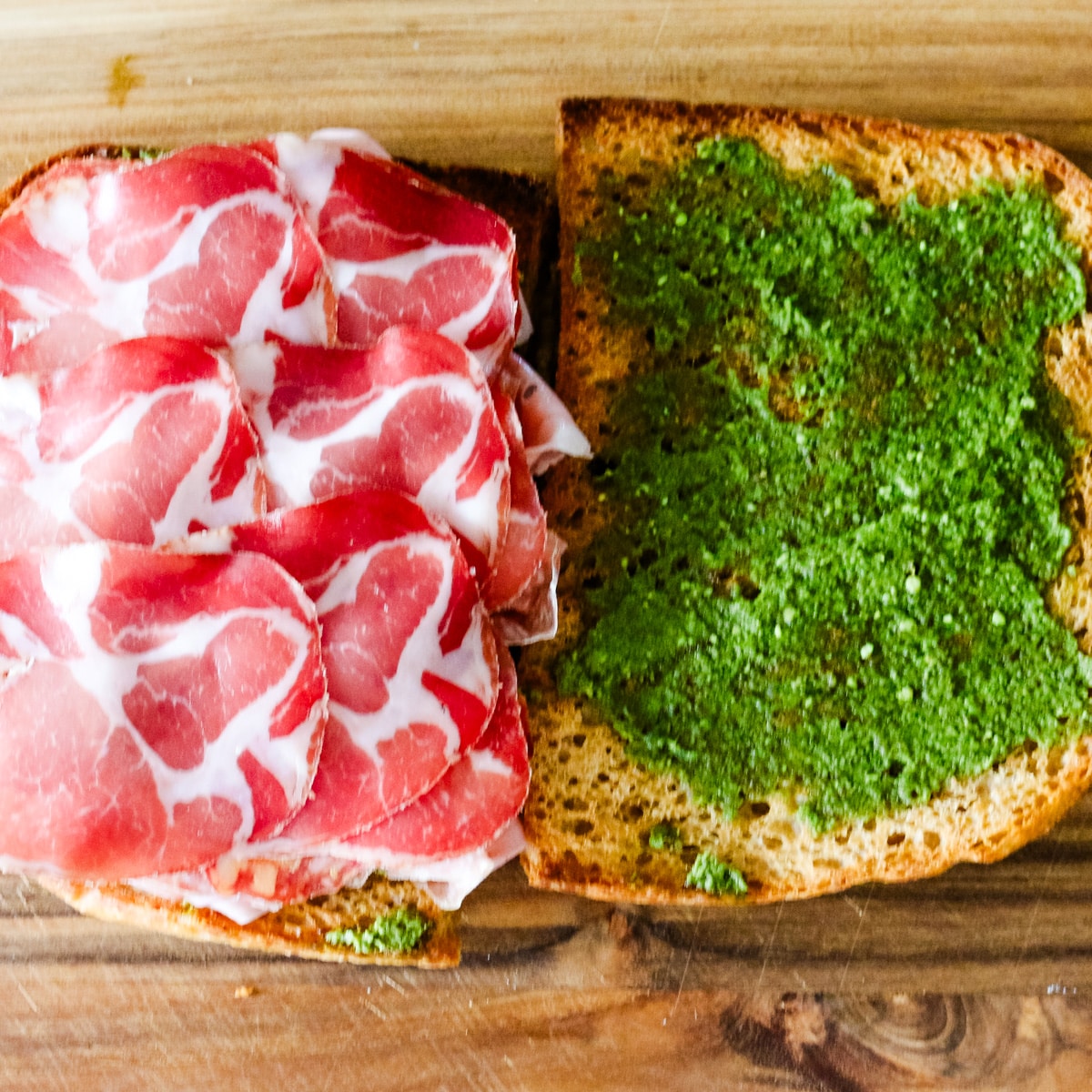 Toasted whole-grain bread slices spread with vibrant green pesto and layered with thinly sliced marbled cured meat on a wooden cutting board.