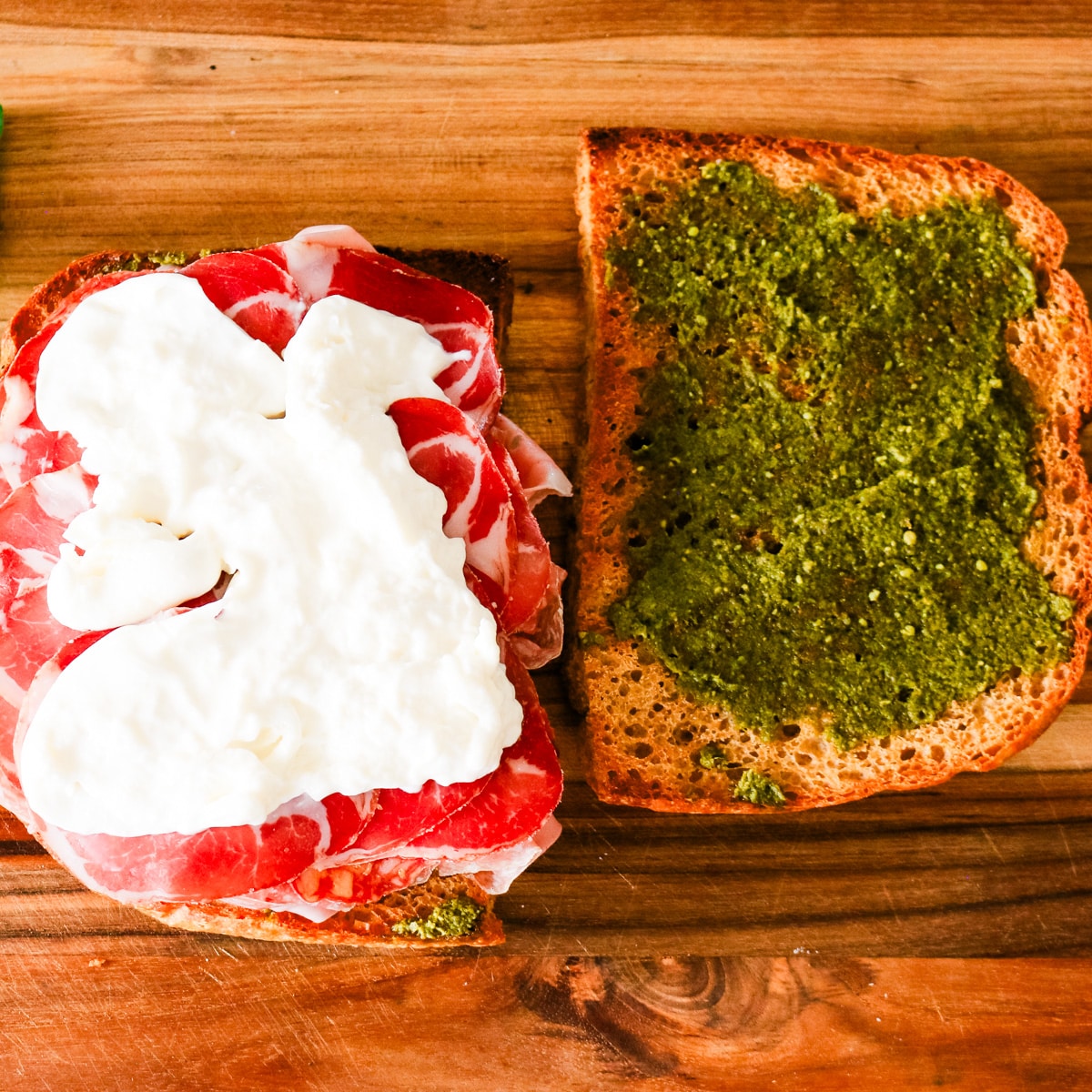 A top-down view of sliced focaccia bread on a wooden board, with fresh mozzarella spread over layered cured meats on one slice, next to a top slice coated in green pesto.