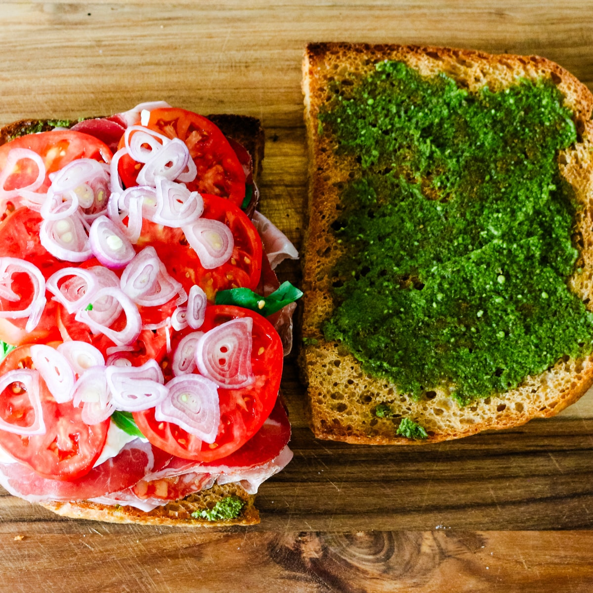 Assembling an Italian sandwich, showing one slice of focaccia loaded with cured meats, ripe tomato slices, and thinly sliced shallots, next to a second slice spread with pesto.