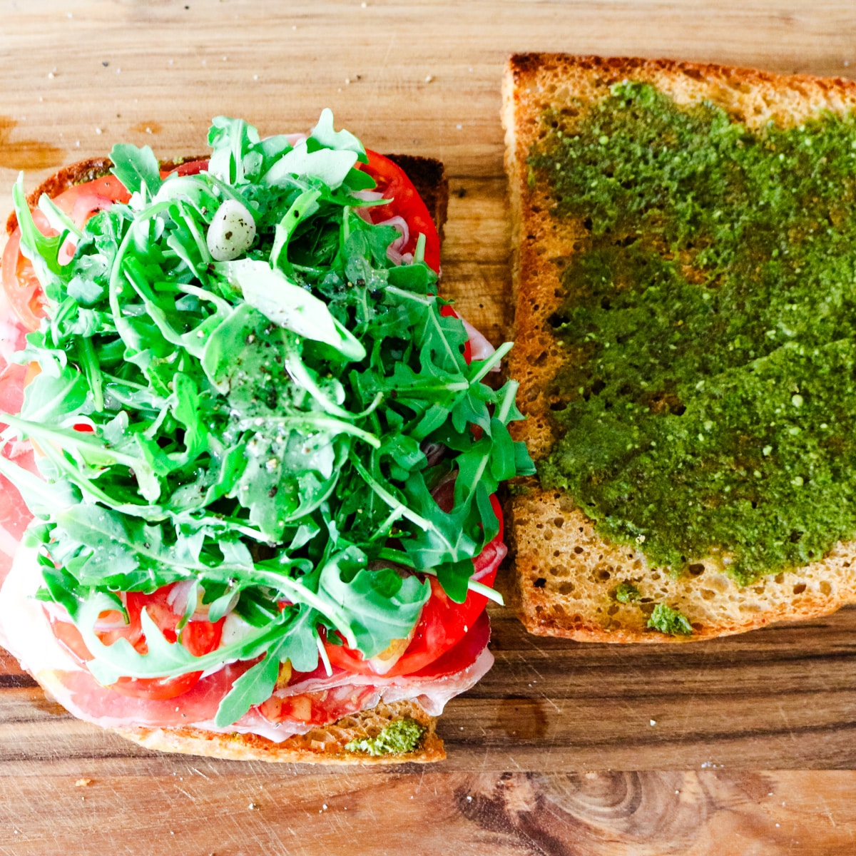 A top-down view of an open-faced focaccia sandwich on a wooden board, with fresh arugula and sliced tomatoes layered over cured meats on one slice, and basil pesto spread evenly across the top slice.