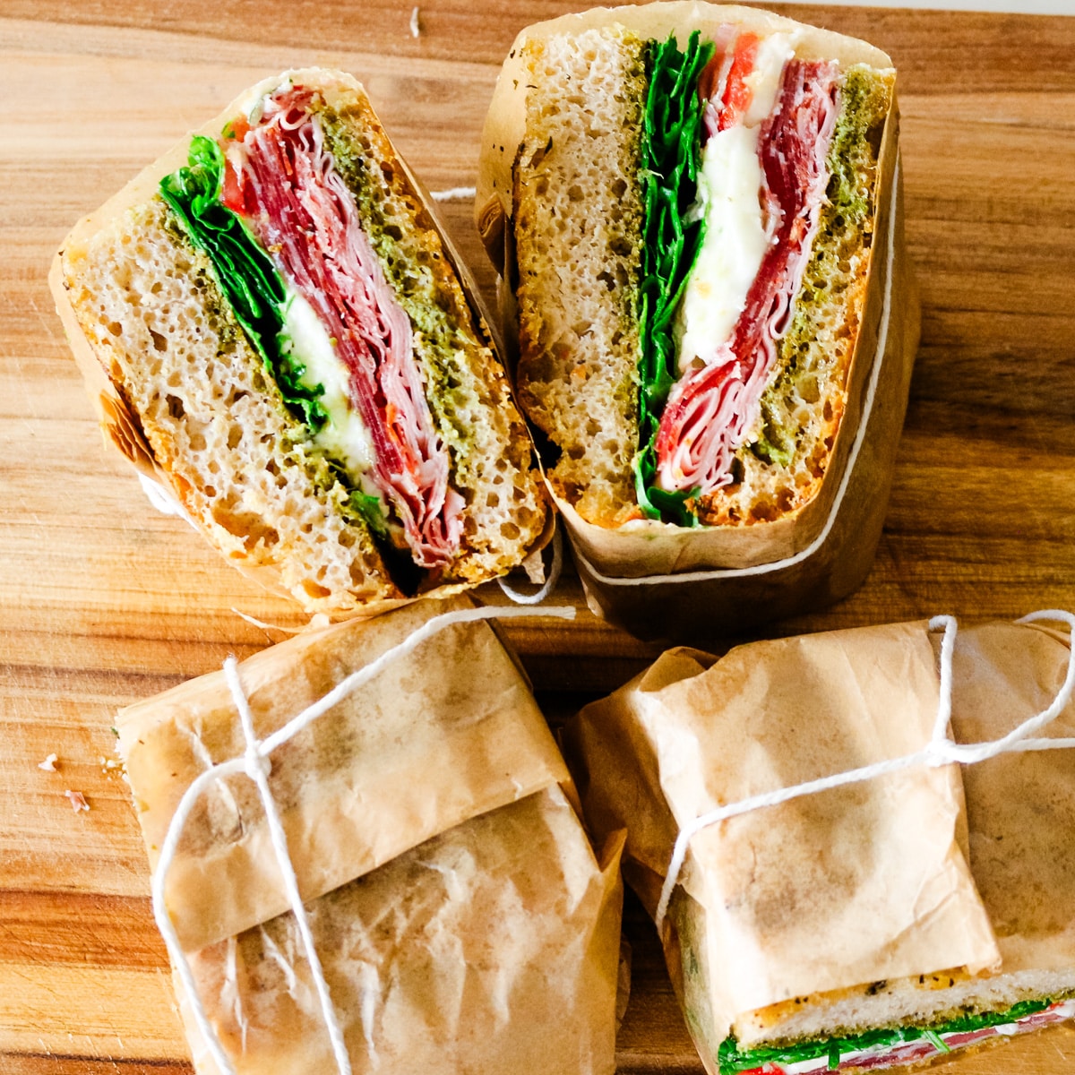 Italian sandwiches wrapped in parchment paper and tied with string, cut into halves to reveal cross-sections of cured meats, fresh mozzarella, pesto, and greens.