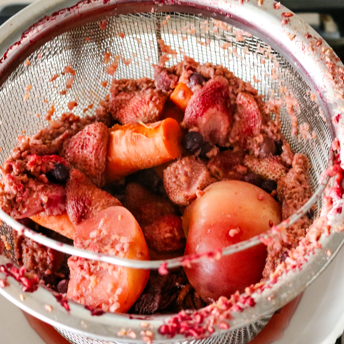 Straining the cooked fruit mixture through a stainless steel mesh colander to yield clear kompot juice.