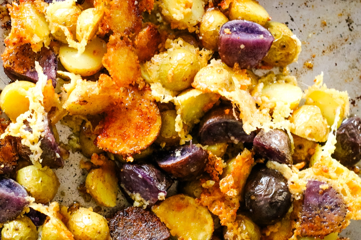 Cooked baby potatoes spread across a metal baking sheet, covered in melted, crispy parmesan cheese.