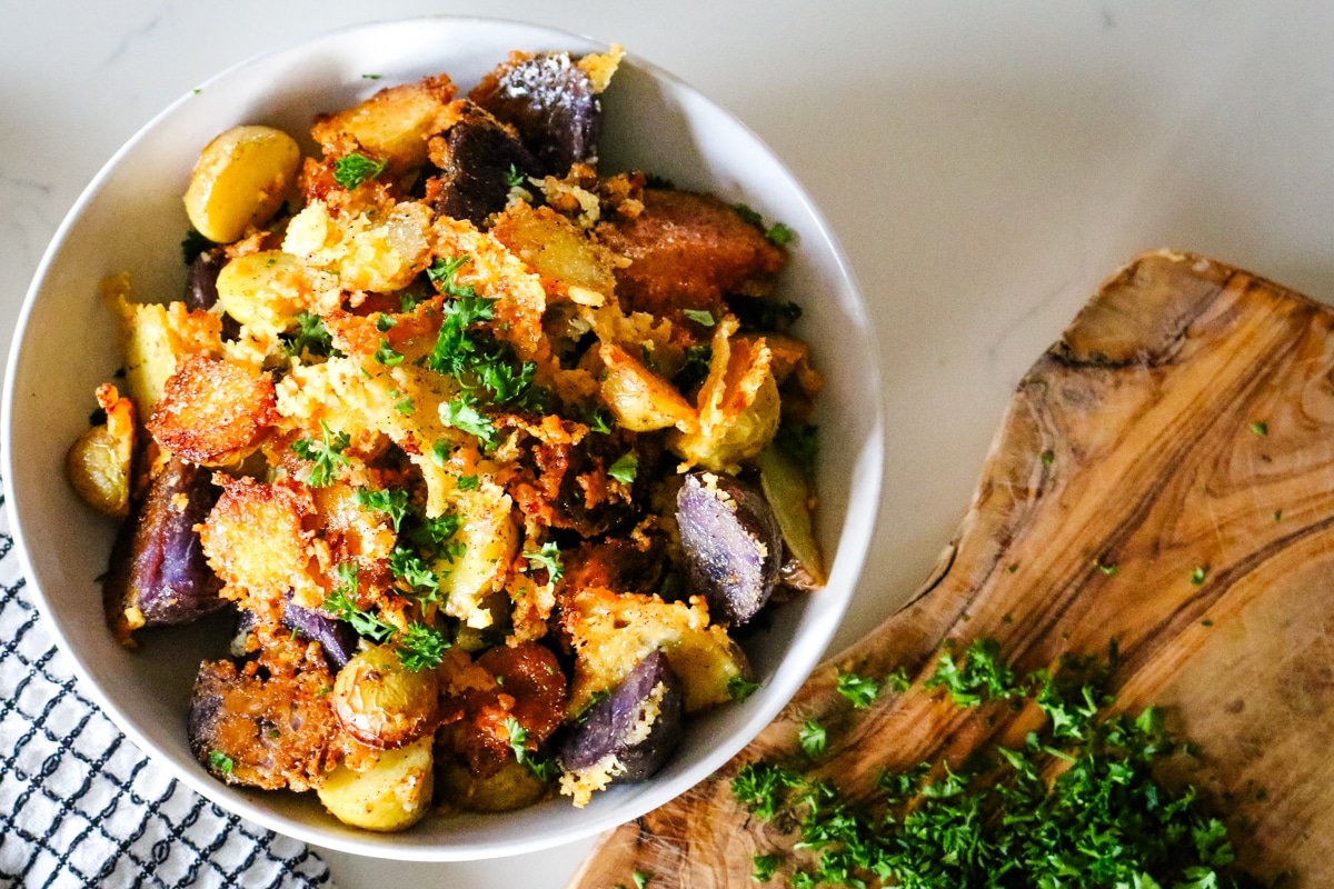 Parmesan crusted potatoes in a bowl topped with extra crispy cheese, next to a cutting board with fresh parsley.