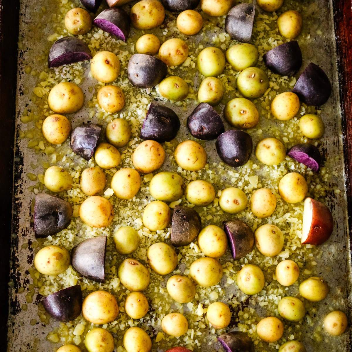 Raw baby potatoes tossed with oil and grated parmesan cheese on a metal baking sheet, prepared for roasting.