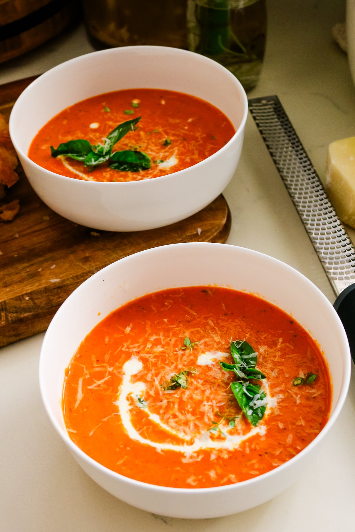Vertical view of two bowls of homemade tomato bisque next to a wooden cutting board and cheese grater.