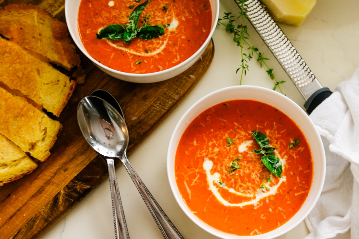 Two white bowls of creamy tomato bisque garnished with fresh basil and parmesan next to sourdough grilled cheese and a cheese grater.