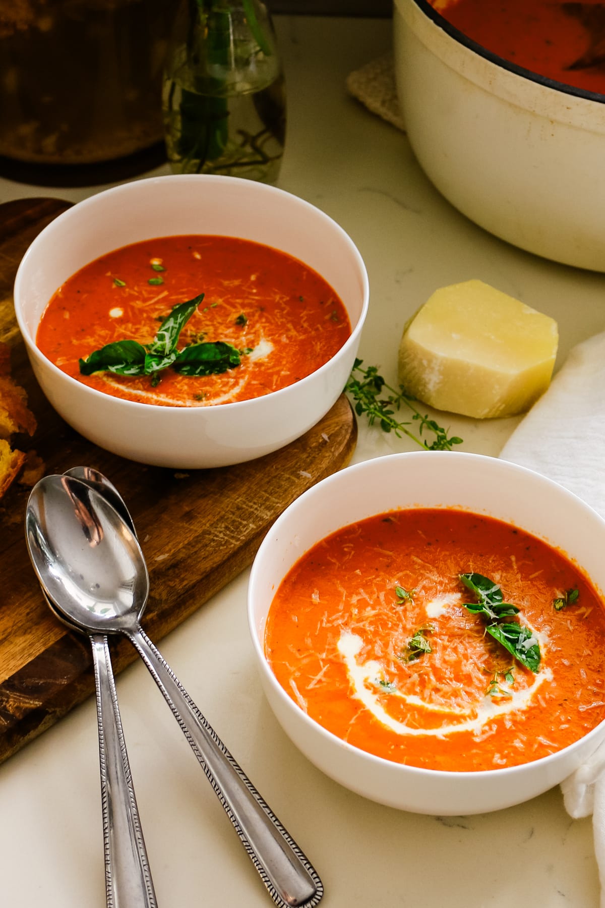 Two white bowls of creamy tomato bisque garnished with fresh basil, cream, and parmesan cheese.