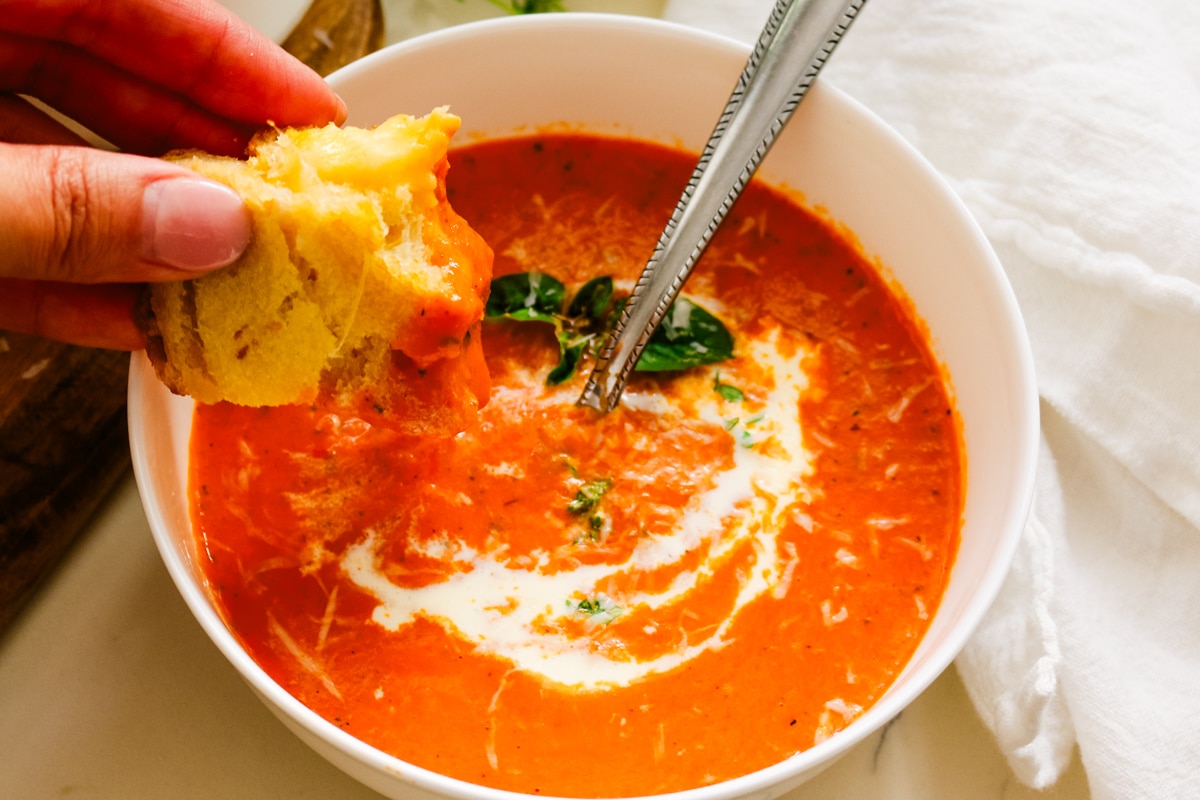 Hand dipping a piece of melted sourdough grilled cheese into a bowl of creamy tomato bisque topped with cream and basil.