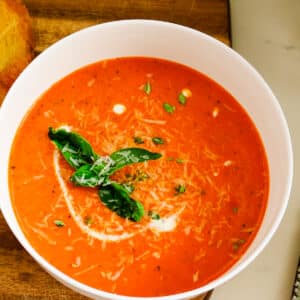 A bowl of homemade tomato bisque topped with fresh basil leaves and shredded parmesan cheese next to grilled sourdough.