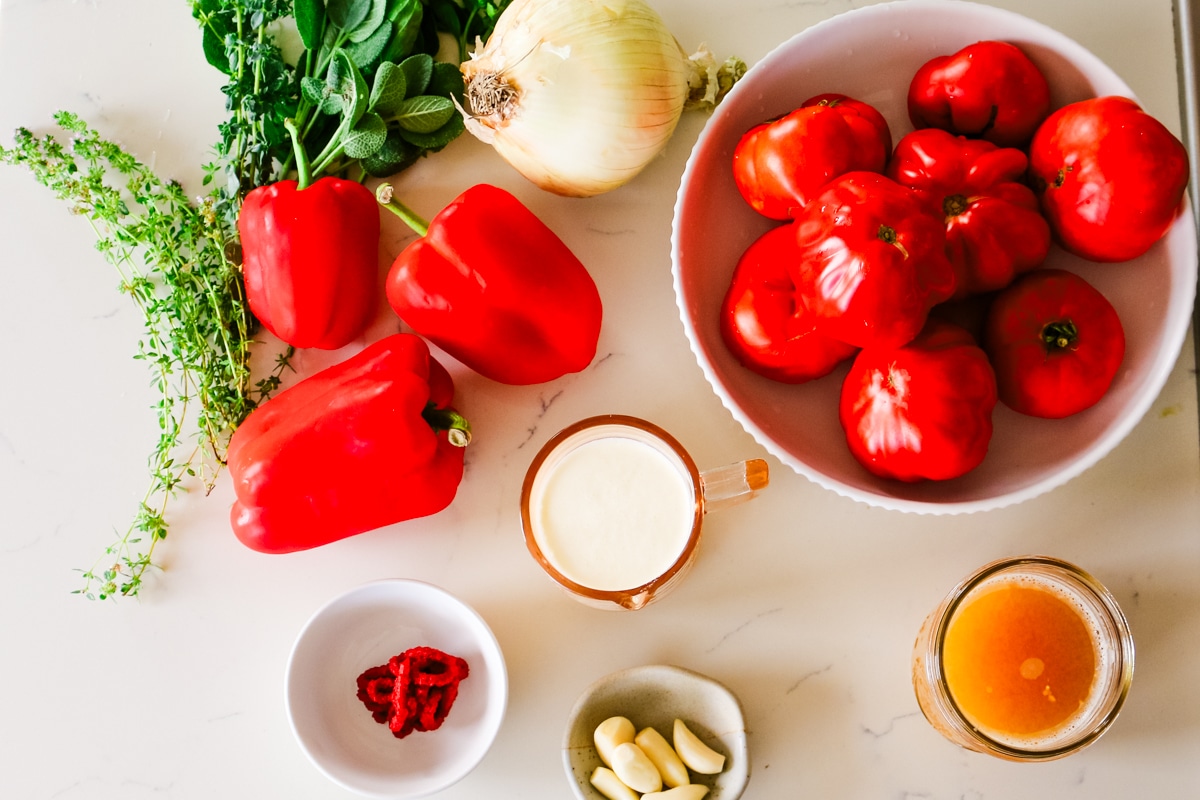Fresh tomatoes, red bell peppers, onion, herbs, cream, and broth prepped on a countertop for roasted tomato bisque.