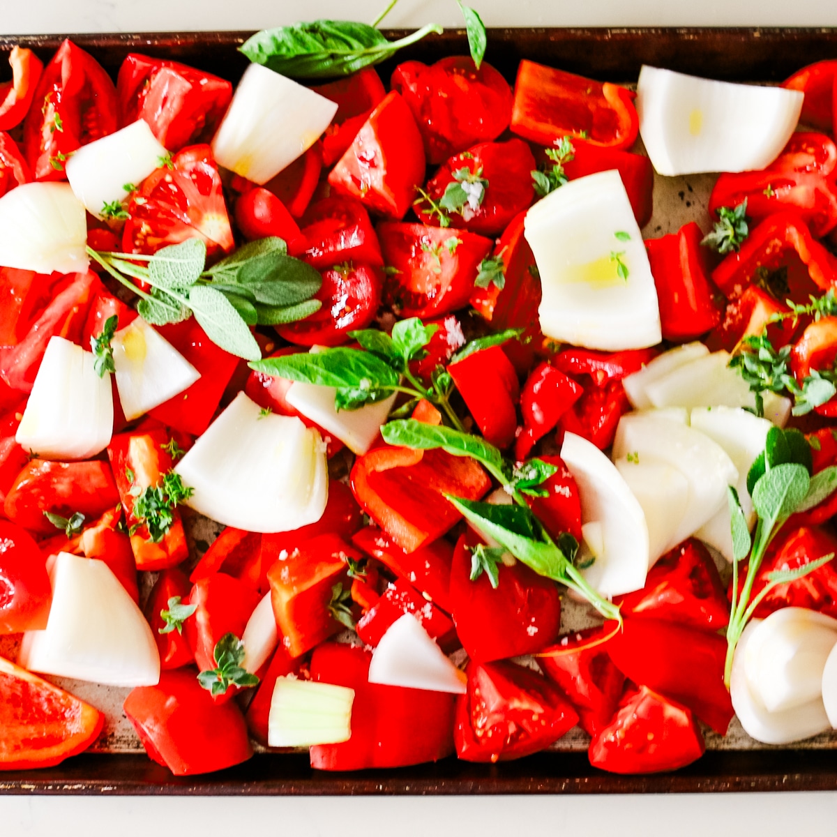 Freshly chopped tomatoes, white onions, red bell peppers, and herbs spread on a baking sheet before roasting for tomato bisque.