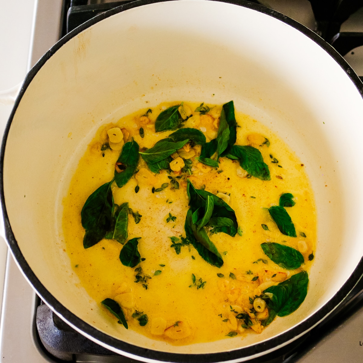 Sliced garlic, fresh basil leaves, and thyme leaves sizzling in melted butter inside a white Dutch oven.