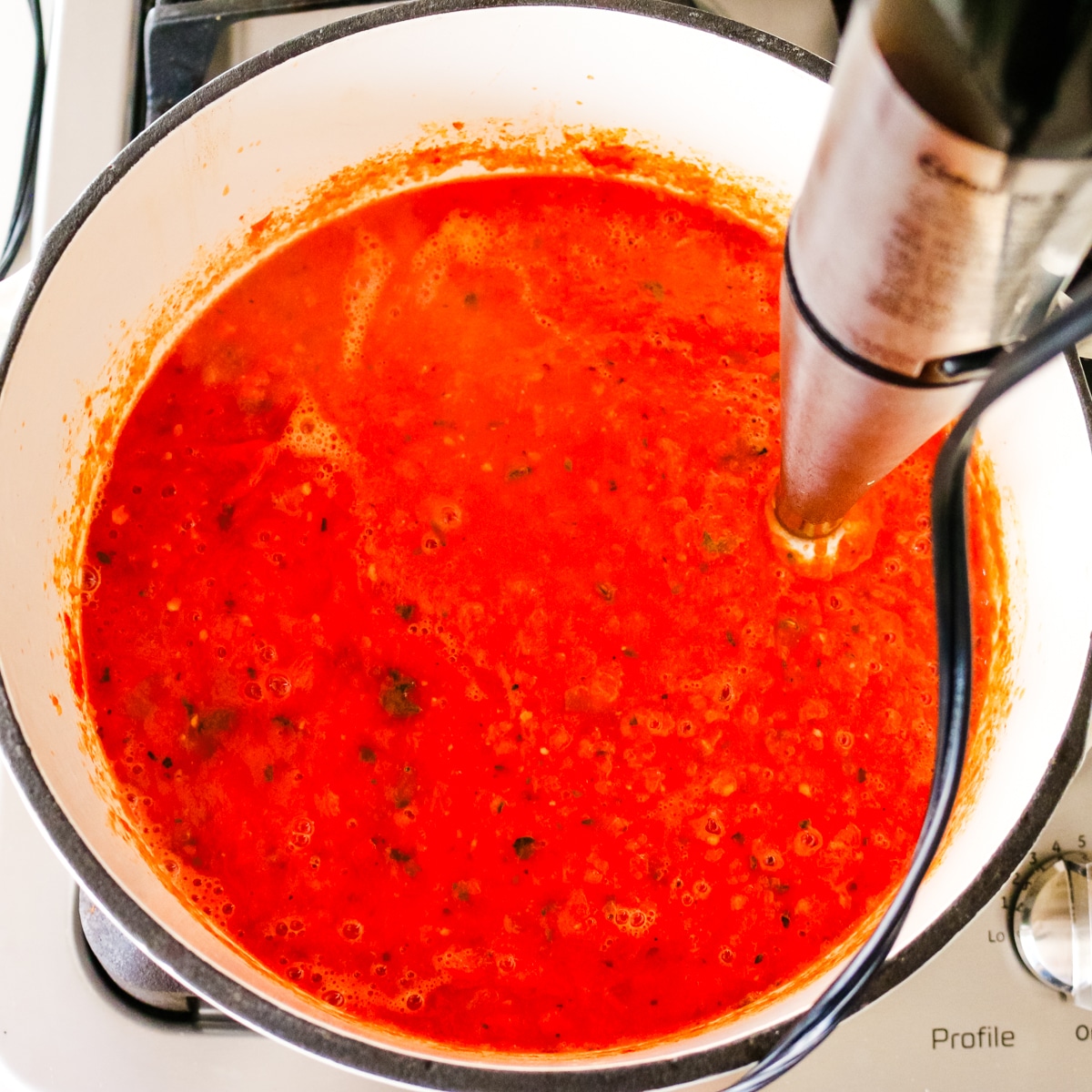 Immersion blender puréeing tomato bisque directly inside a white Dutch oven on the stove.