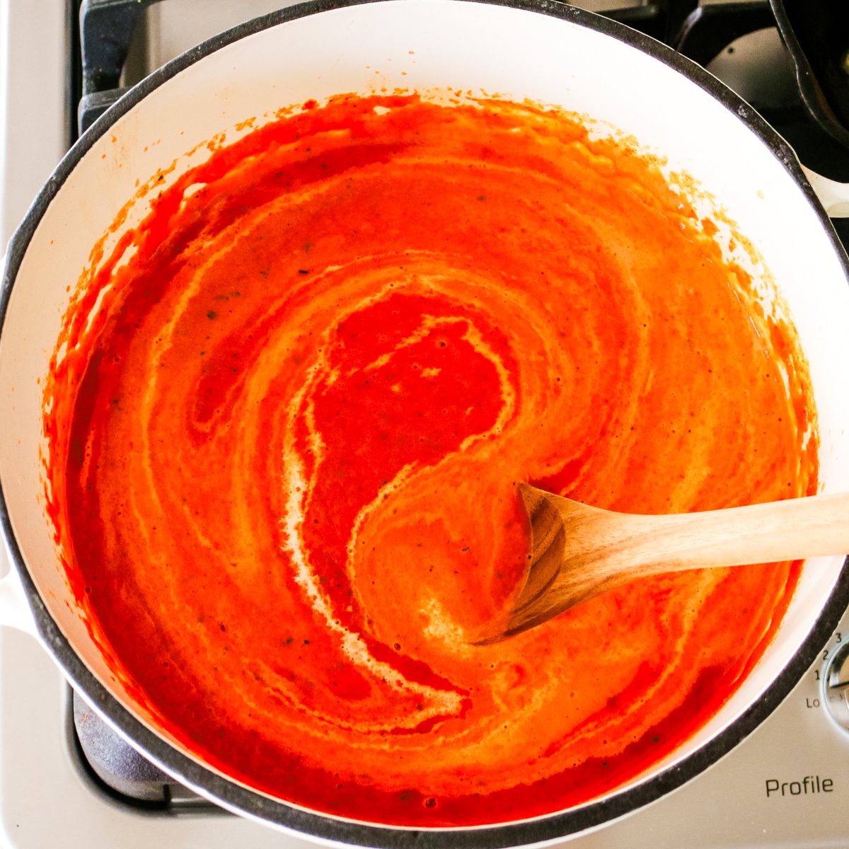 Heavy cream being stirred with a wooden spoon into pureed tomato bisque in a white Dutch oven.