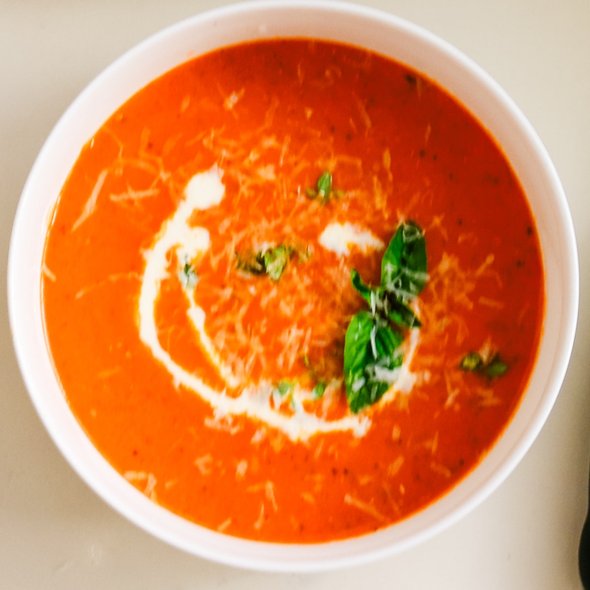 A white ceramic bowl filled with rich homemade tomato bisque topped with fresh basil sprigs and shredded parmesan.