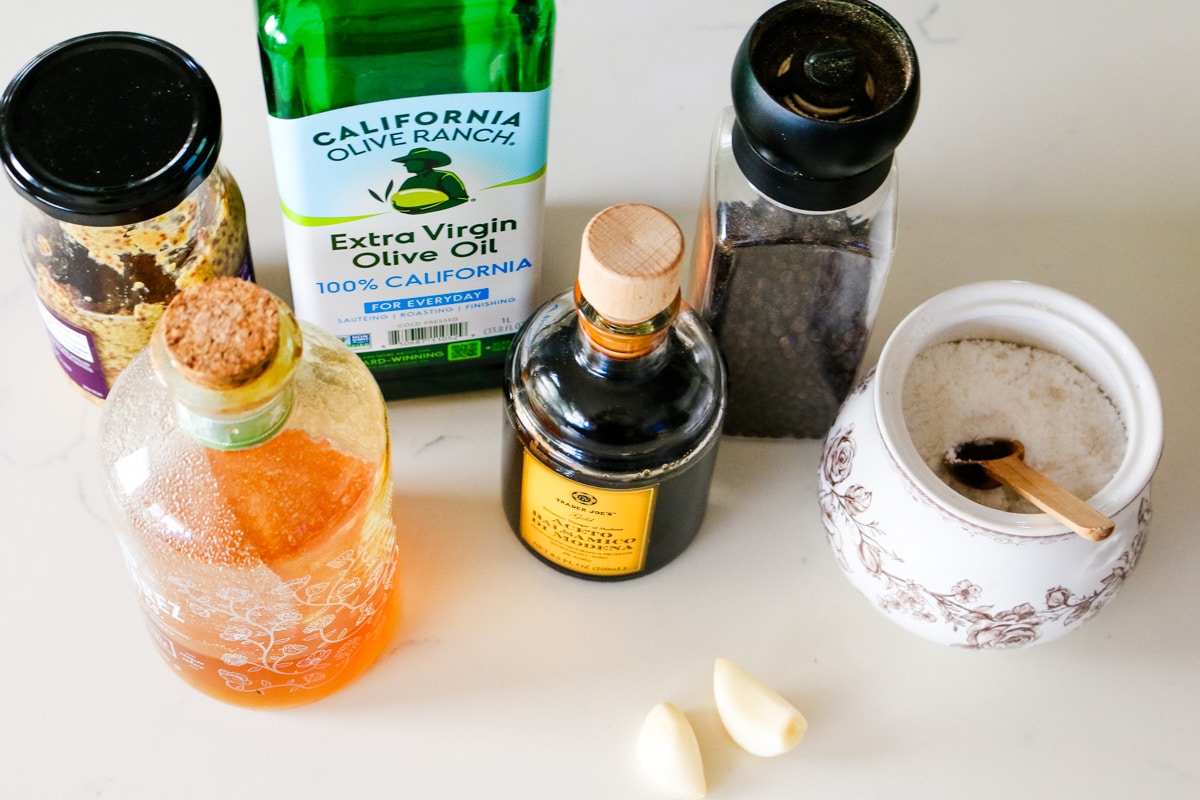 Ingredients for homemade balsamic vinaigrette arranged on a white counter, including olive oil, balsamic vinegar, honey, grain mustard, garlic, salt, and pepper.