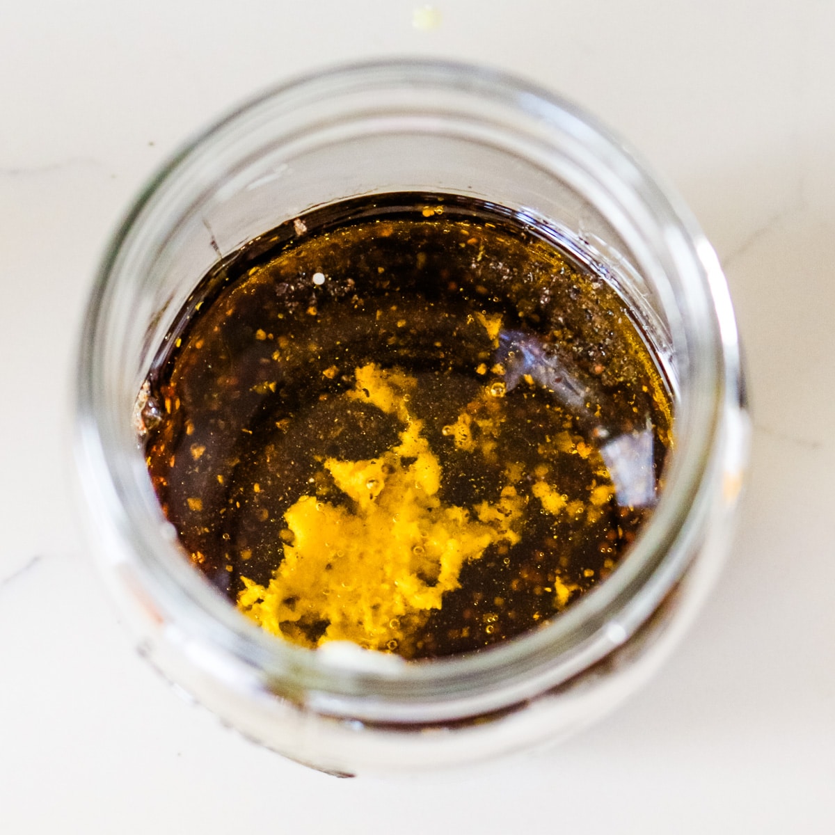 Looking down into a glass jar containing olive oil, balsamic vinegar, minced garlic, and seasonings before mixing.