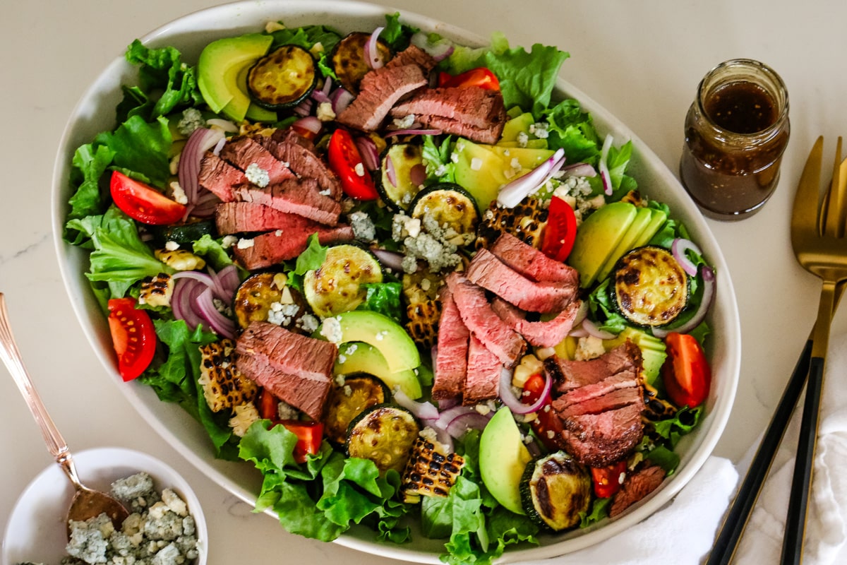 steak salad in a white bowl with a jar of balsamic vinaigrette on the side and a bowl of blue cheese.